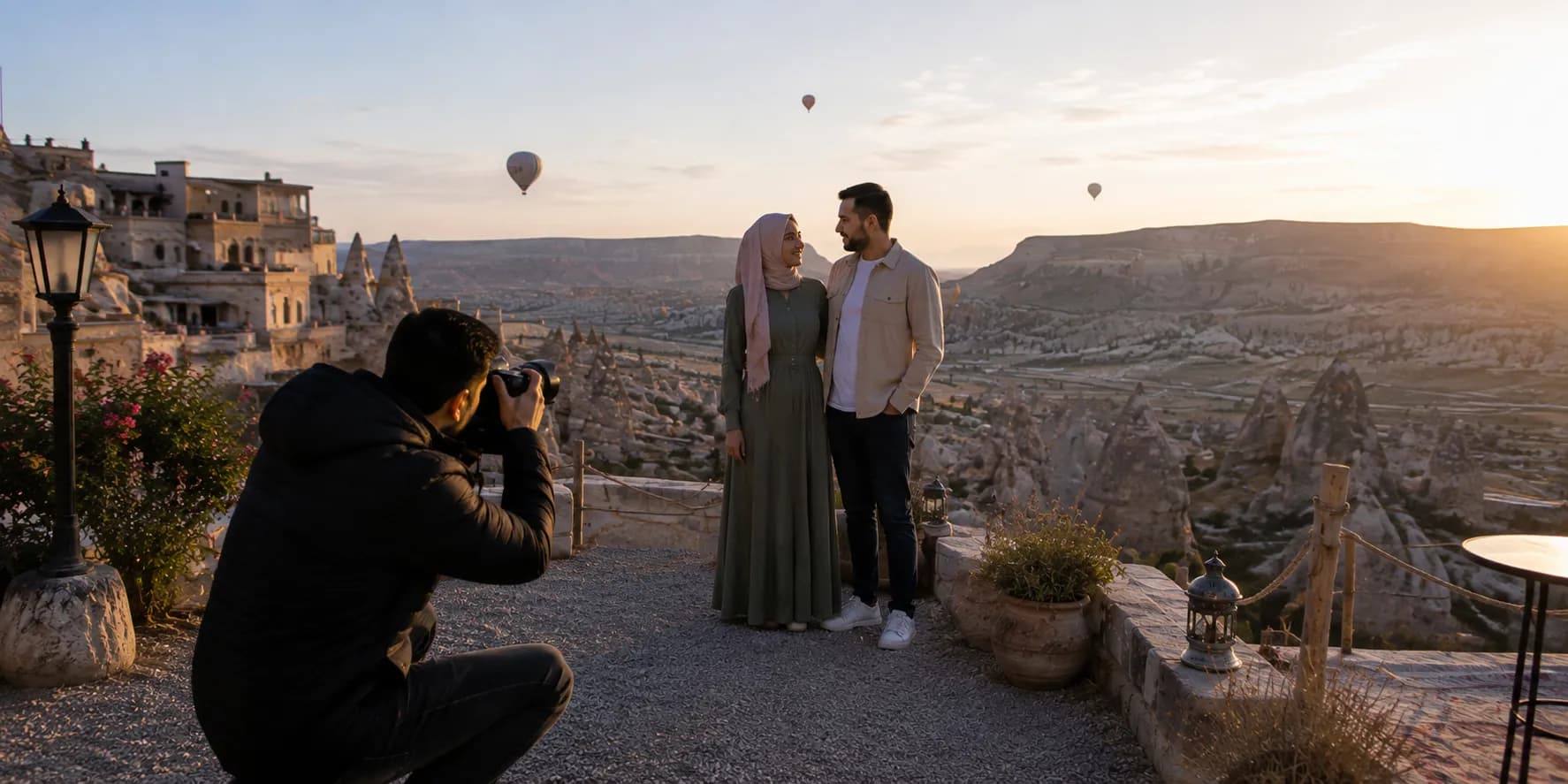 A photographer shooting a couple on a Cappadocian terrace at sunrise, hot air balloons behind them