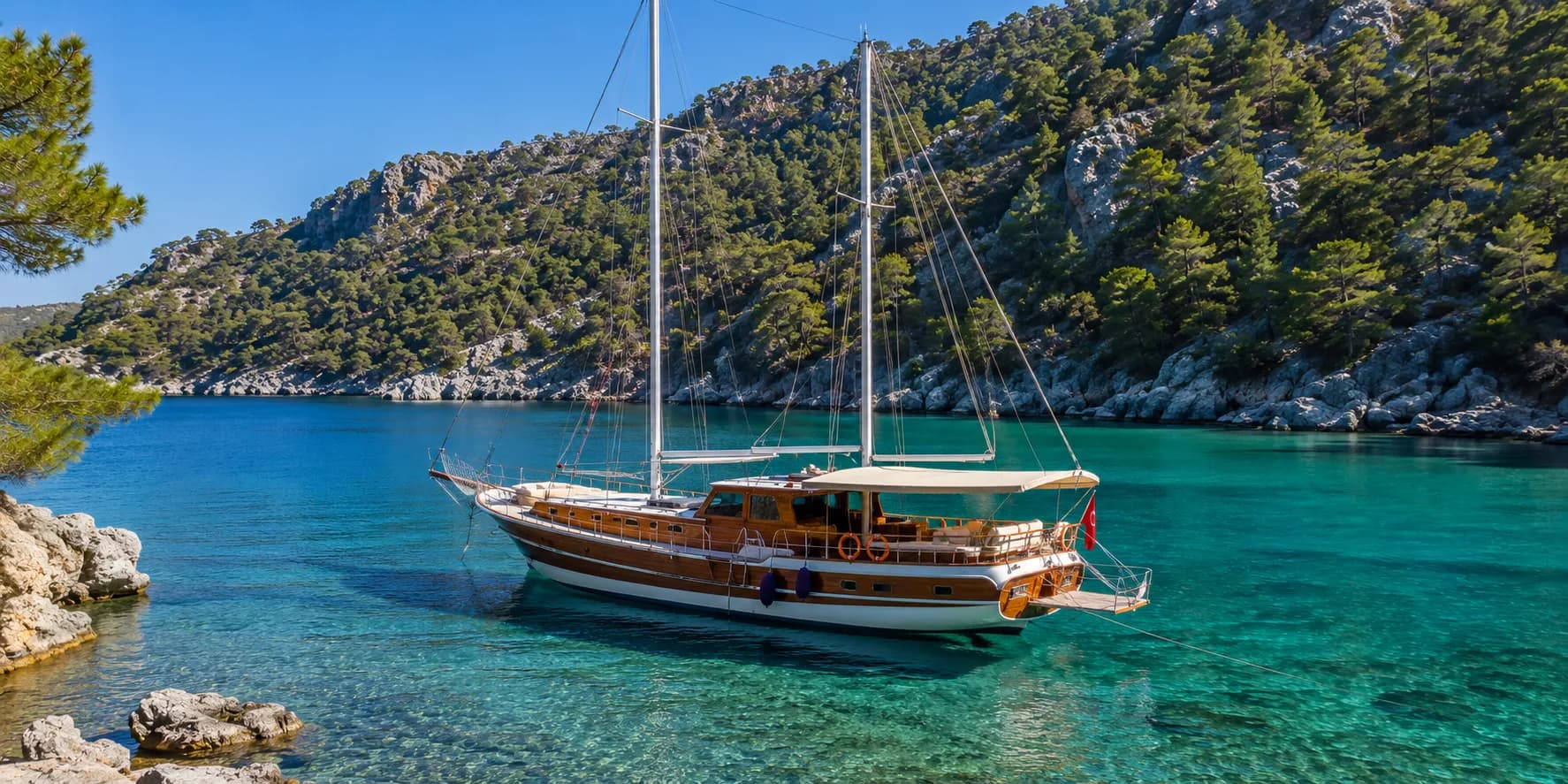 A traditional wooden Turkish gulet anchored in a clear turquoise bay below pine-covered hills